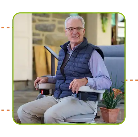 An older man with gray hair and glasses sits on a motorized stairlift chair indoors, smiling. A potted plant is nearby.